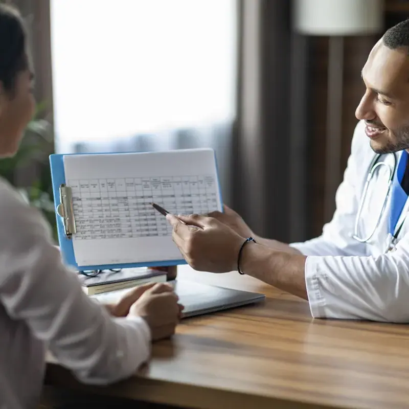 A physician pointing to a results chart on a clipboard while a patient looks on across a consultation desk