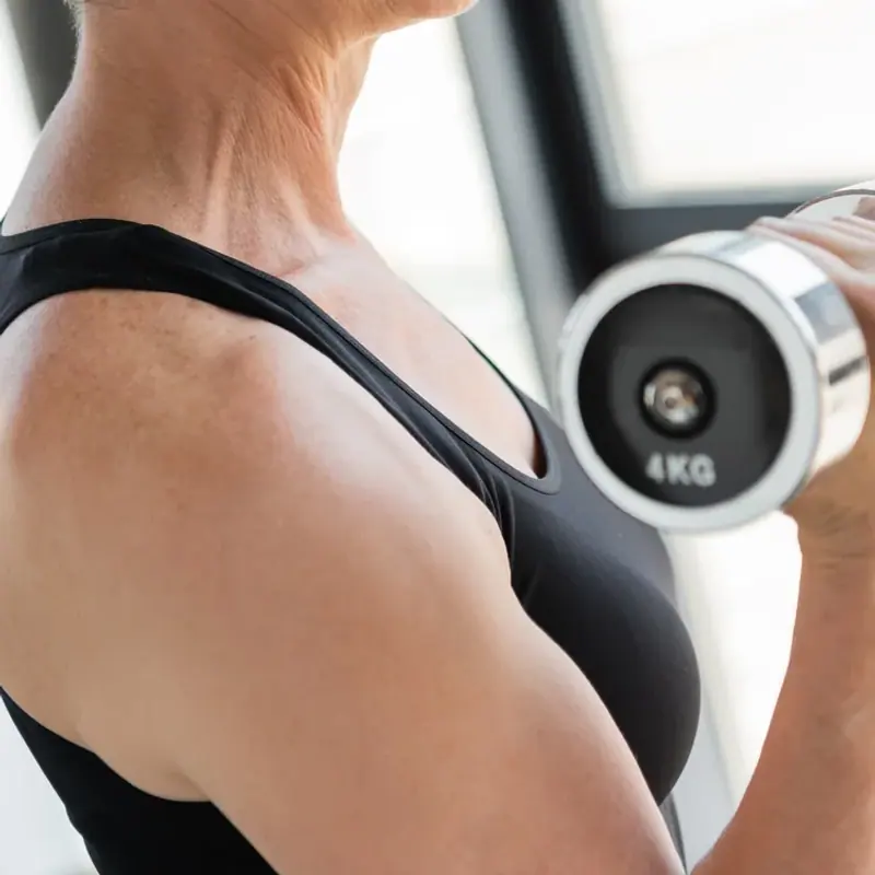 A woman in her sixties lifting a dumbbell beside a window in a bright gym