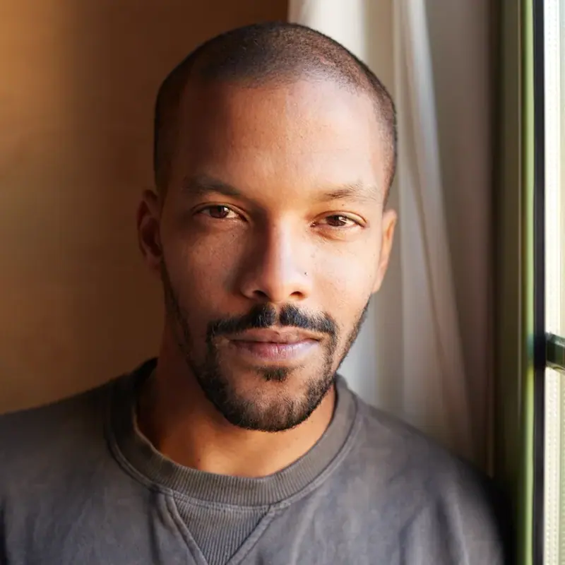 A man in a grey sweatshirt beside a window, lit by warm daylight, looking at the camera