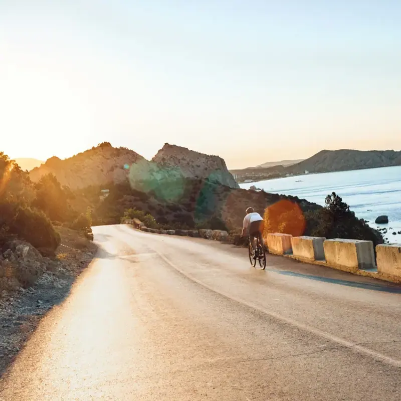A cyclist riding a winding coastal road beside the sea in low evening sun