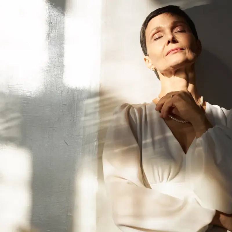 A woman in a white dress standing in warm window light, eyes closed, hand at her collar