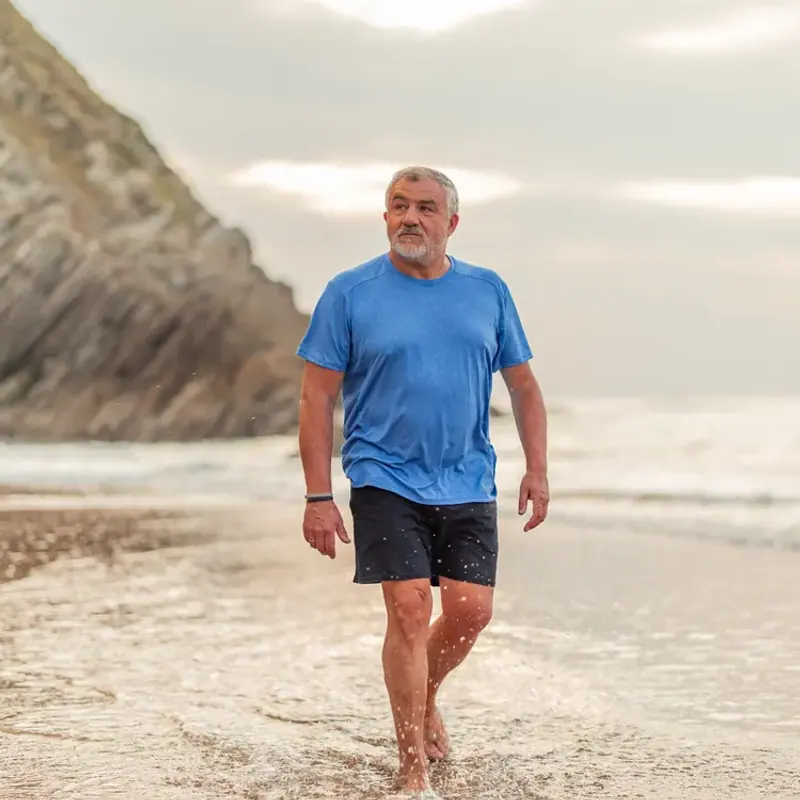 An older man walking barefoot along the water’s edge beneath a rocky cliff