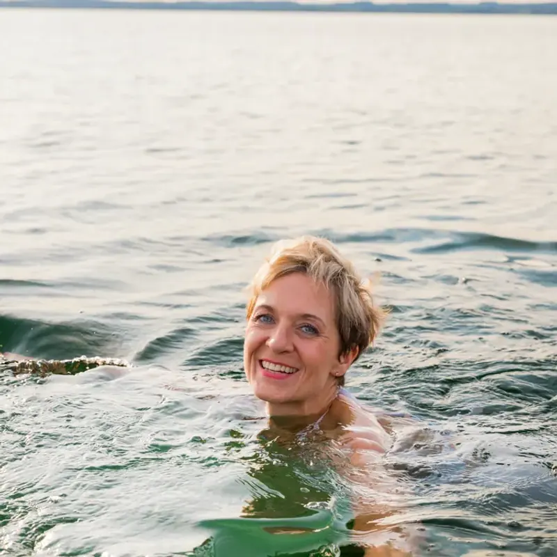 A woman with short blond hair smiling as she swims in a wide, calm lake