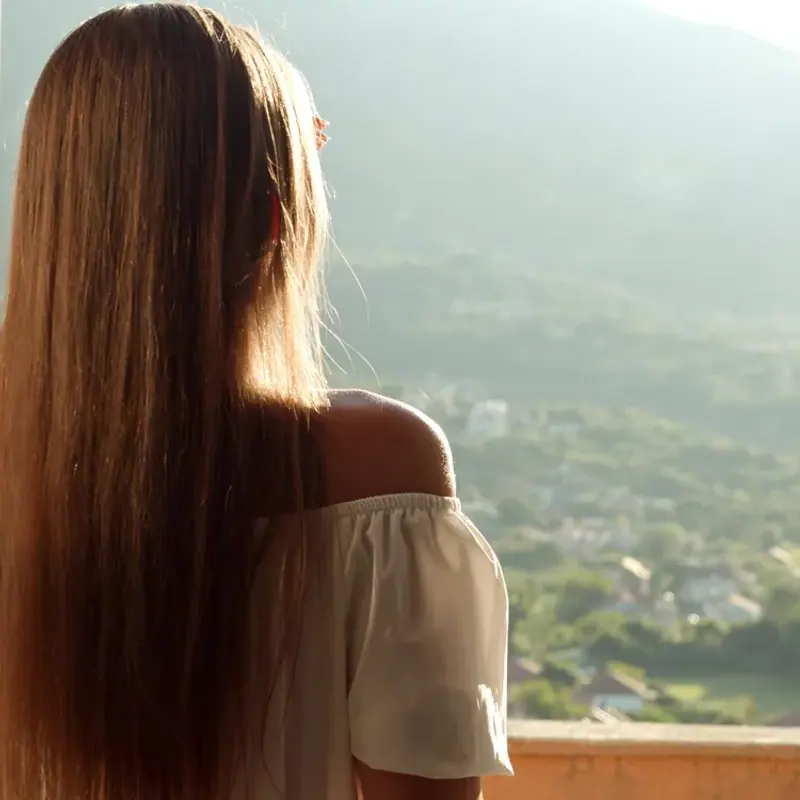 A woman with long brown hair at an open shuttered window overlooking green hills