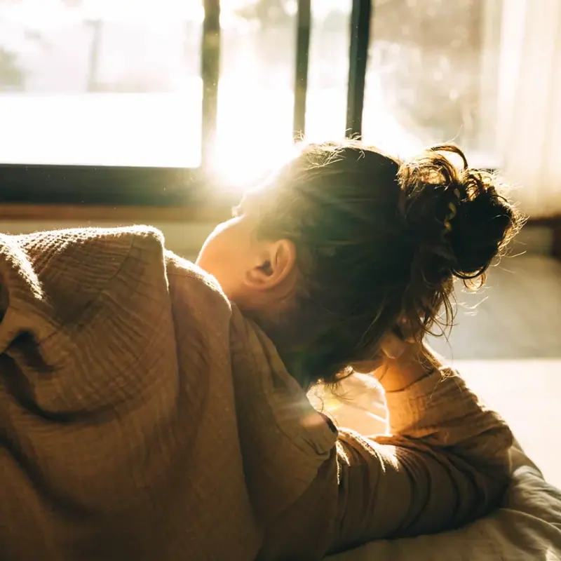 A woman resting her head on her arms in bed, backlit by low morning sun
