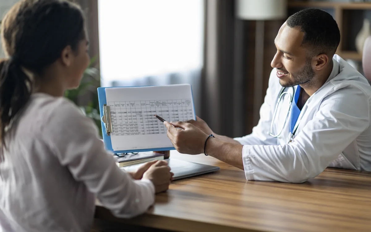 A physician pointing to a results chart on a clipboard while a patient looks on across a consultation desk