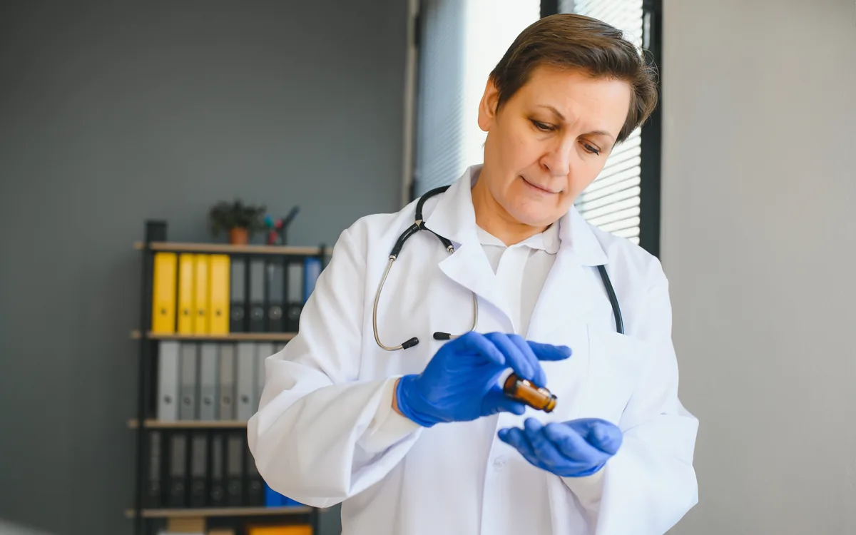 A clinician in a white coat and gloves tipping tablets from a small amber bottle into her gloved hand