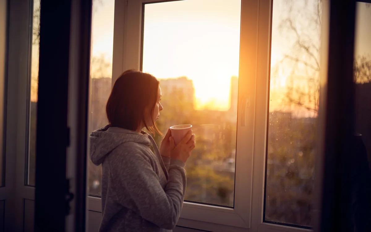 A woman holding a mug at a window, watching the sun come up over rooftops