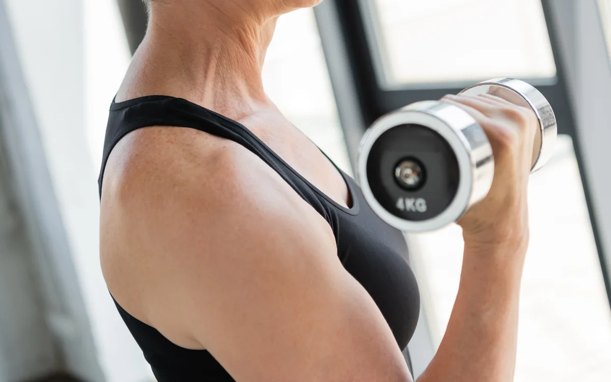 A woman in her sixties lifting a dumbbell beside a window in a bright gym