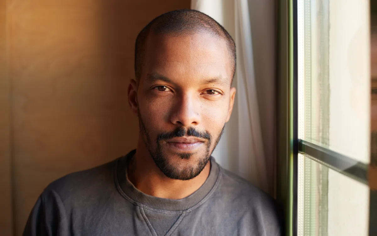 A man in a grey sweatshirt beside a window, lit by warm daylight, looking at the camera