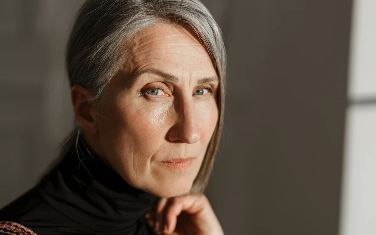 A woman with grey hair in a black turtleneck, photographed in soft indoor light