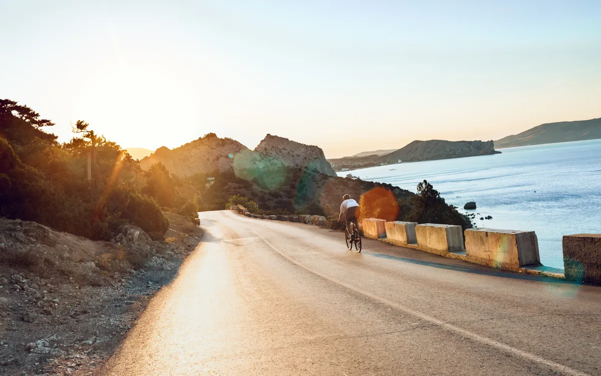 A cyclist riding a winding coastal road beside the sea in low evening sun
