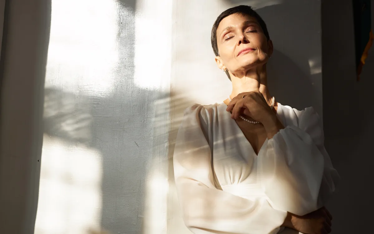 A woman in a white dress standing in warm window light, eyes closed, hand at her collar
